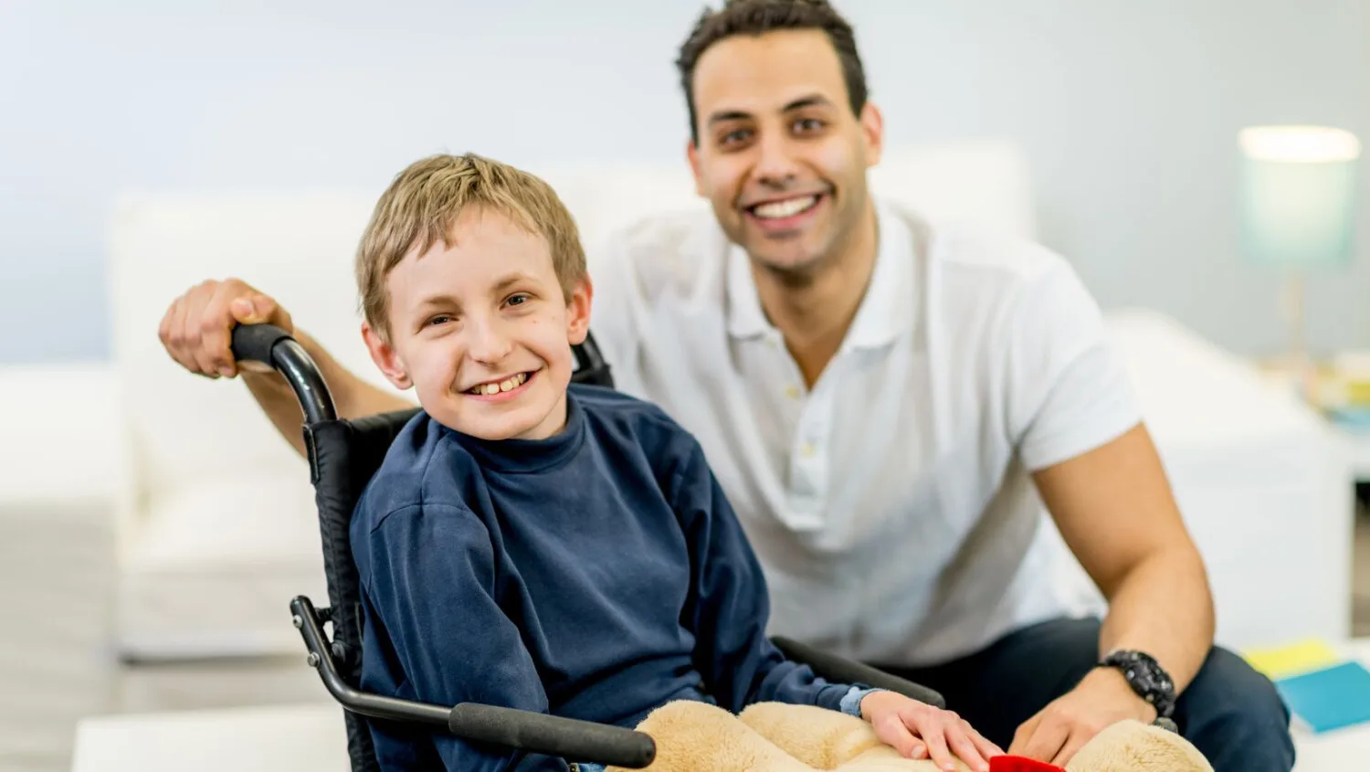 NDIS support worker smiling with young boy in wheelchair during a home disability care session.