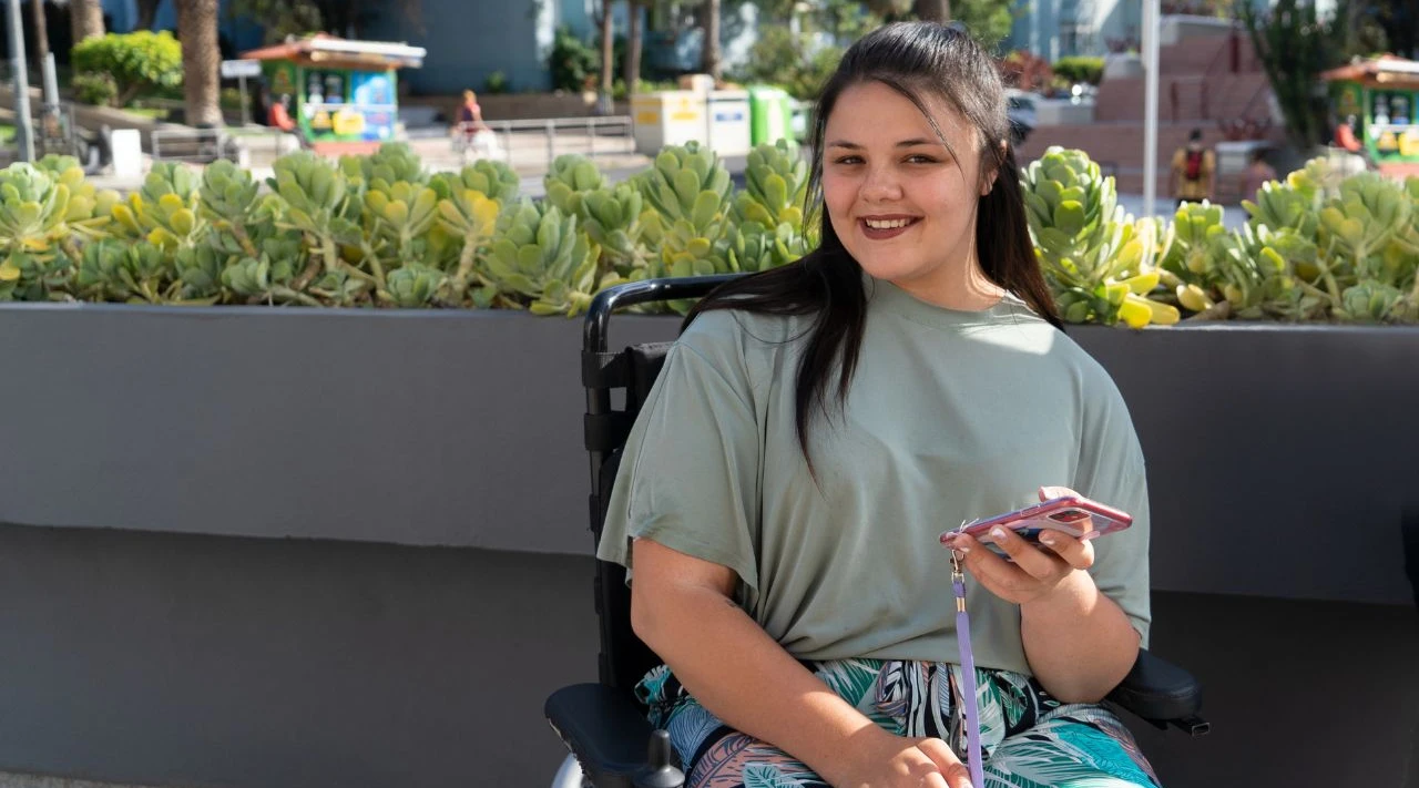 Young woman in a wheelchair smiling outdoors, holding a phone, reflecting independence and confidence.