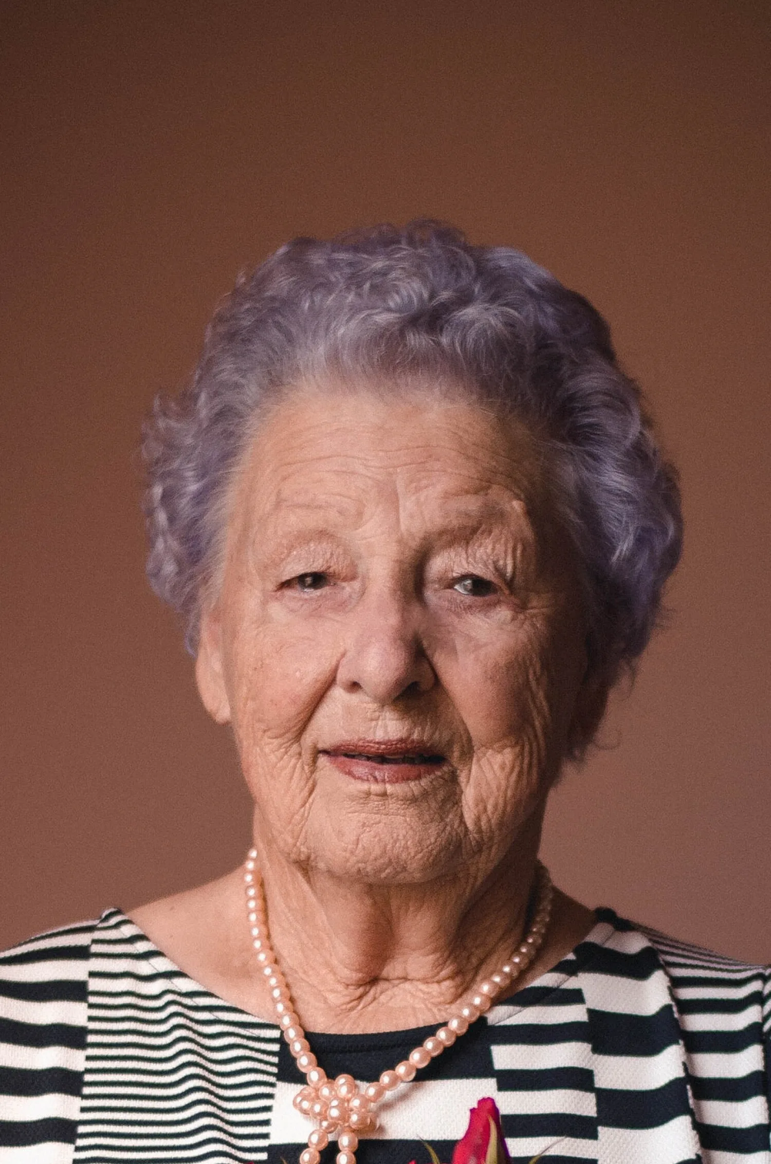 Elderly woman in pearls and stripes cherishes her fresh roses, supported by thoughtful ShiftCare.