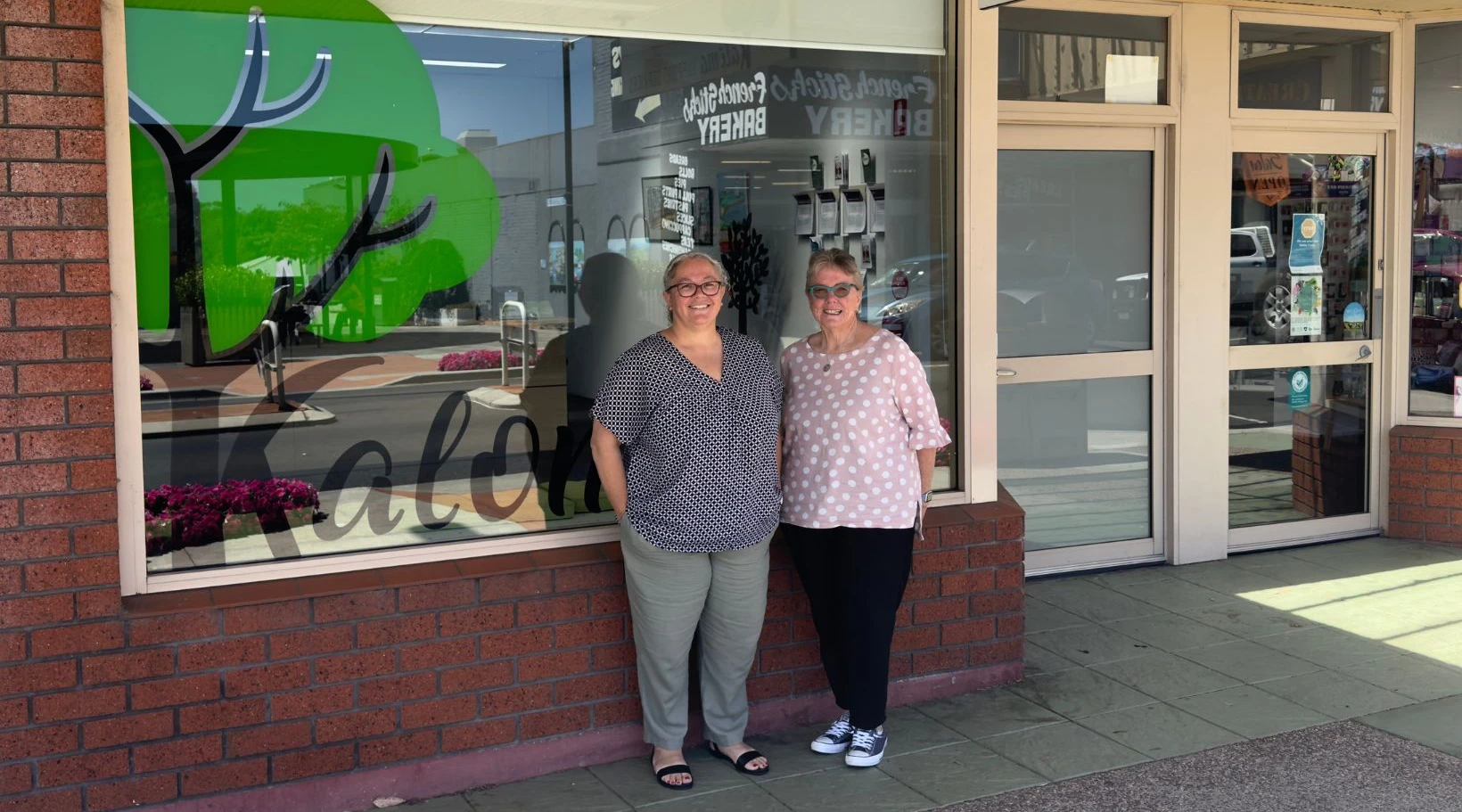 Two smiling women standing outside a local business with glass windows and bright signage.