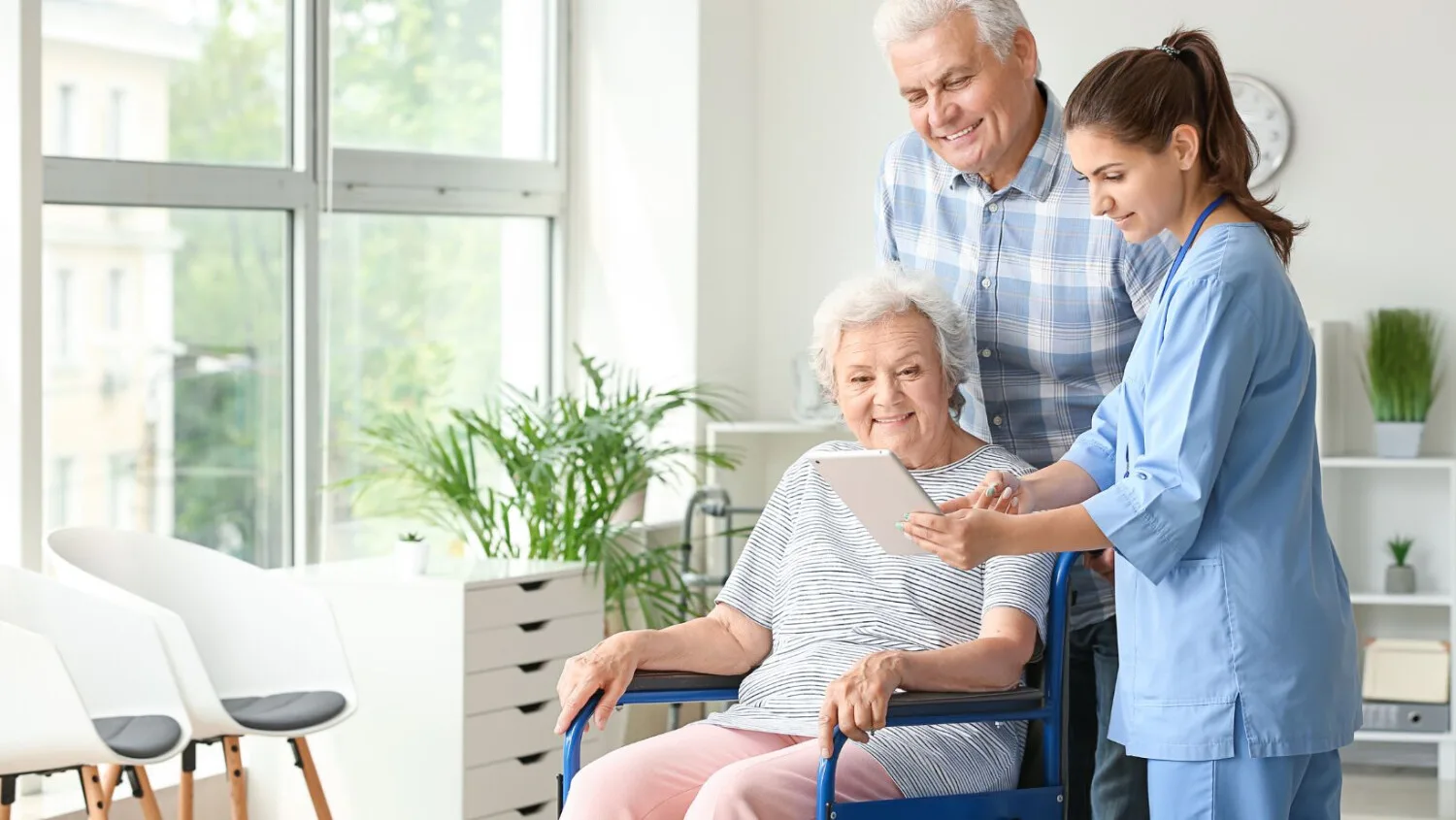 Caregiver showing tablet to senior client in wheelchair with family member during home care visit.