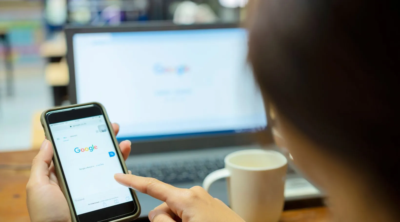 Person searching on Google using a smartphone with a laptop and coffee cup in the background.