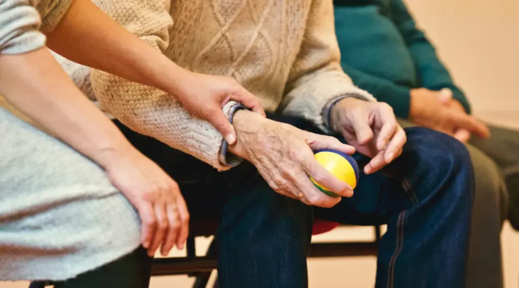 Elderly person holding a therapy ball with caregiver assistance, ShiftCare.