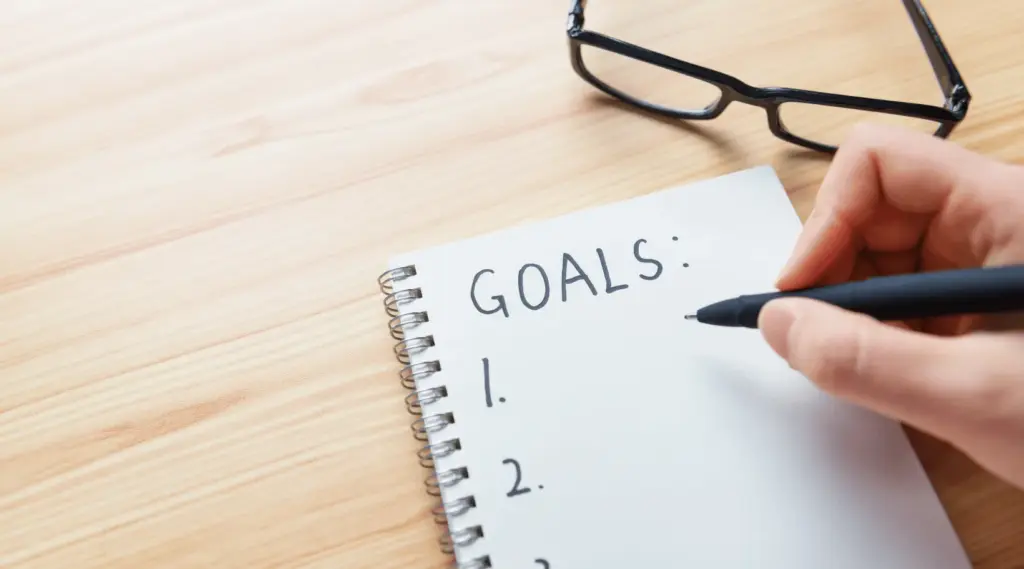 Person writing goals in notebook with pen on desk, symbolizing planning, focus, and motivation.