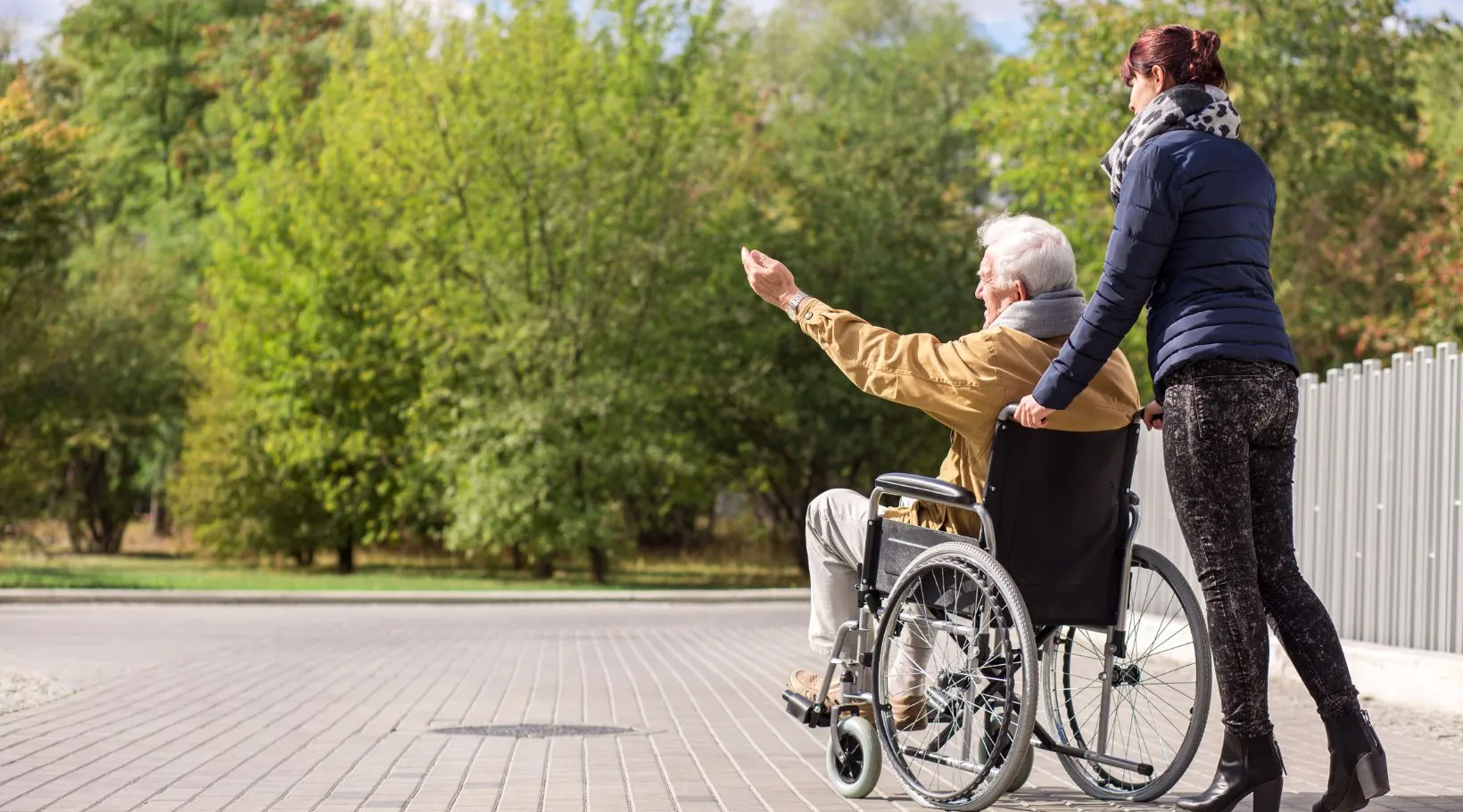 Caregiver walking beside elderly person using a wheelchair in the park, ShiftCare.