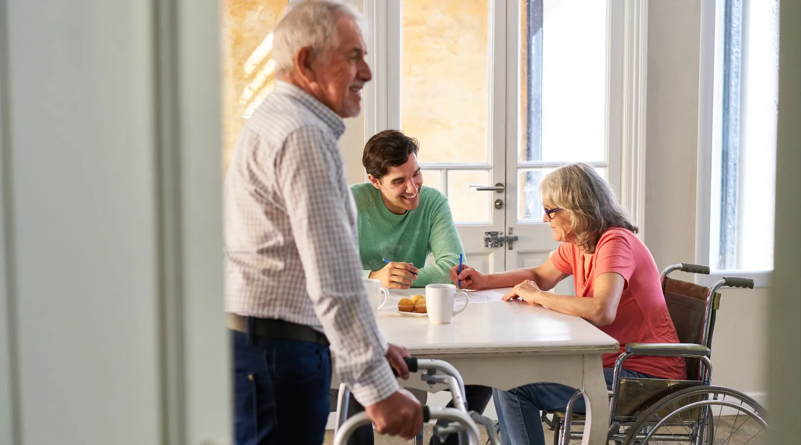 Care provider engaging with elderly man and woman in wheelchair at table, by ShiftCare.