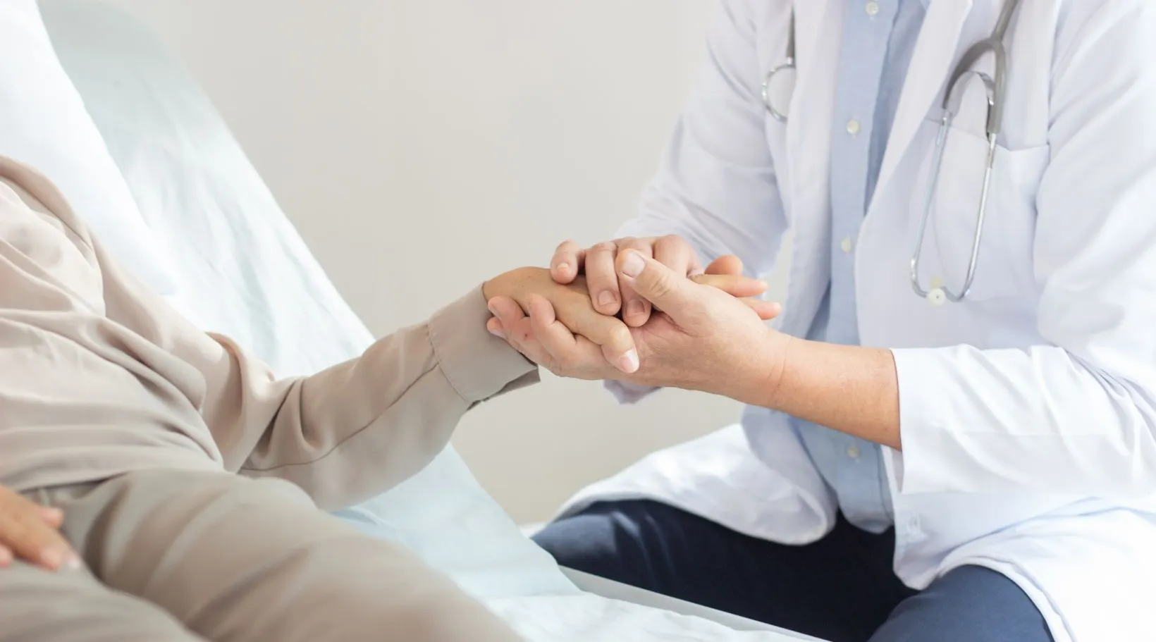 A doctor offers reassurance while holding a patient’s hand during a hospital bedside visit.