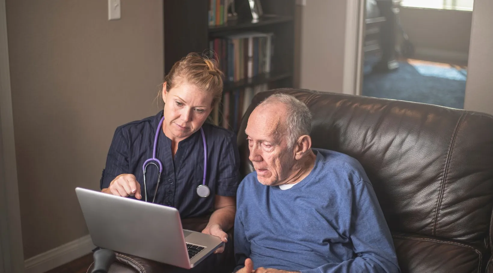 Caregiver showing elderly man health information on laptop at home, ShiftCare.