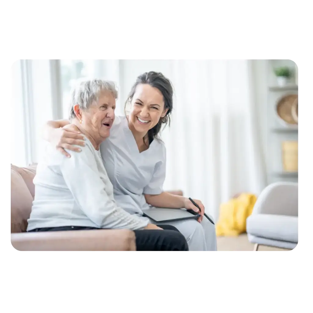 A smiling caregiver embracing an elderly client sitting on a sofa, representing supportive disability care.
