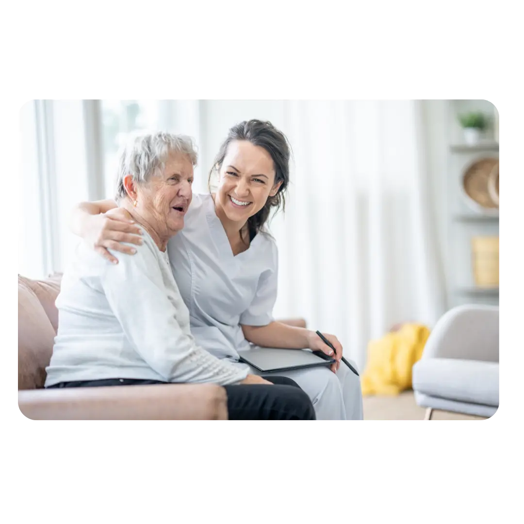 A smiling caregiver embracing an elderly client sitting on a sofa, representing supportive disability care.